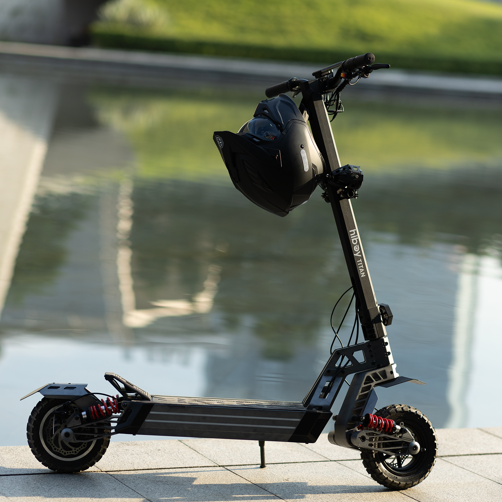 Hiboy TITAN electric scooter parked beside a reflecting pool with a helmet hung on the stem