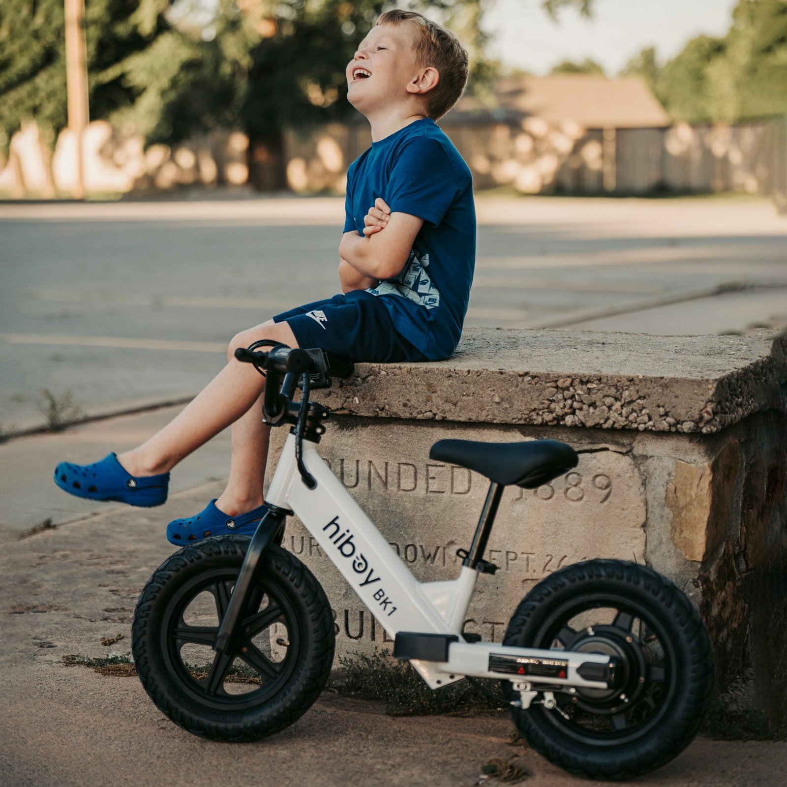 A young child in a helmet sitting on a white Hiboy BK1 electric balance bike beside a wooden park seat