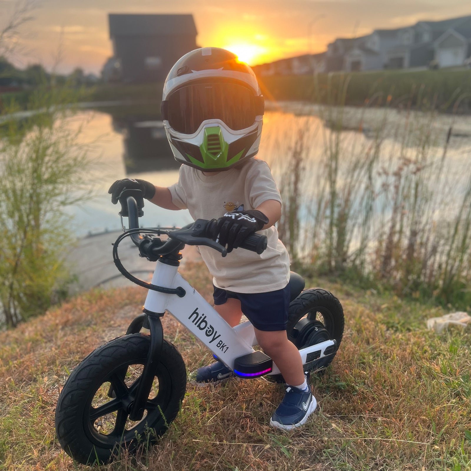 A young child riding a white Hiboy BK1 electric balance bike on a path beside water at sunset