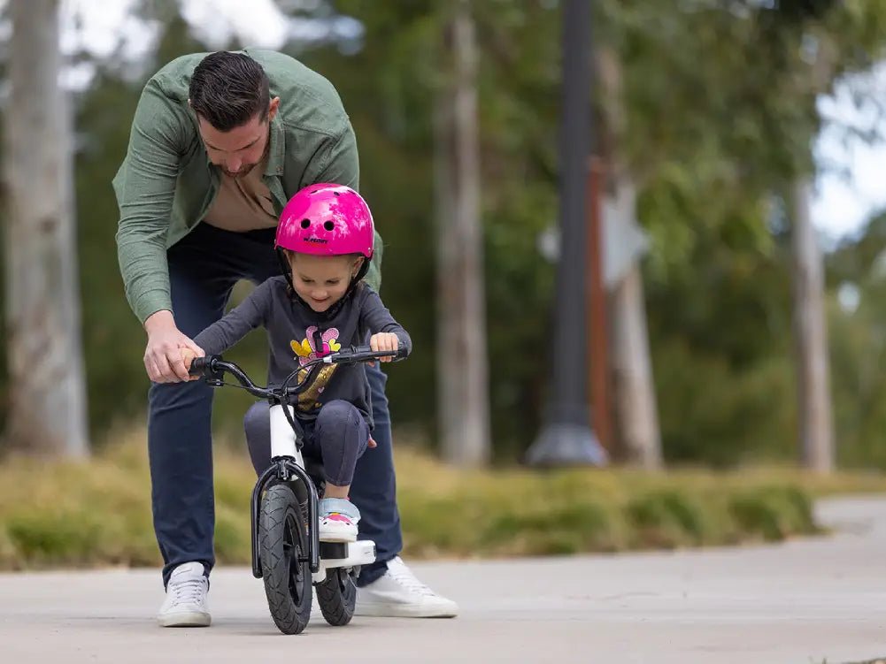 An adult crouching beside a young child in a pink helmet who is seated on a Hiboy BK1 electric balance bike