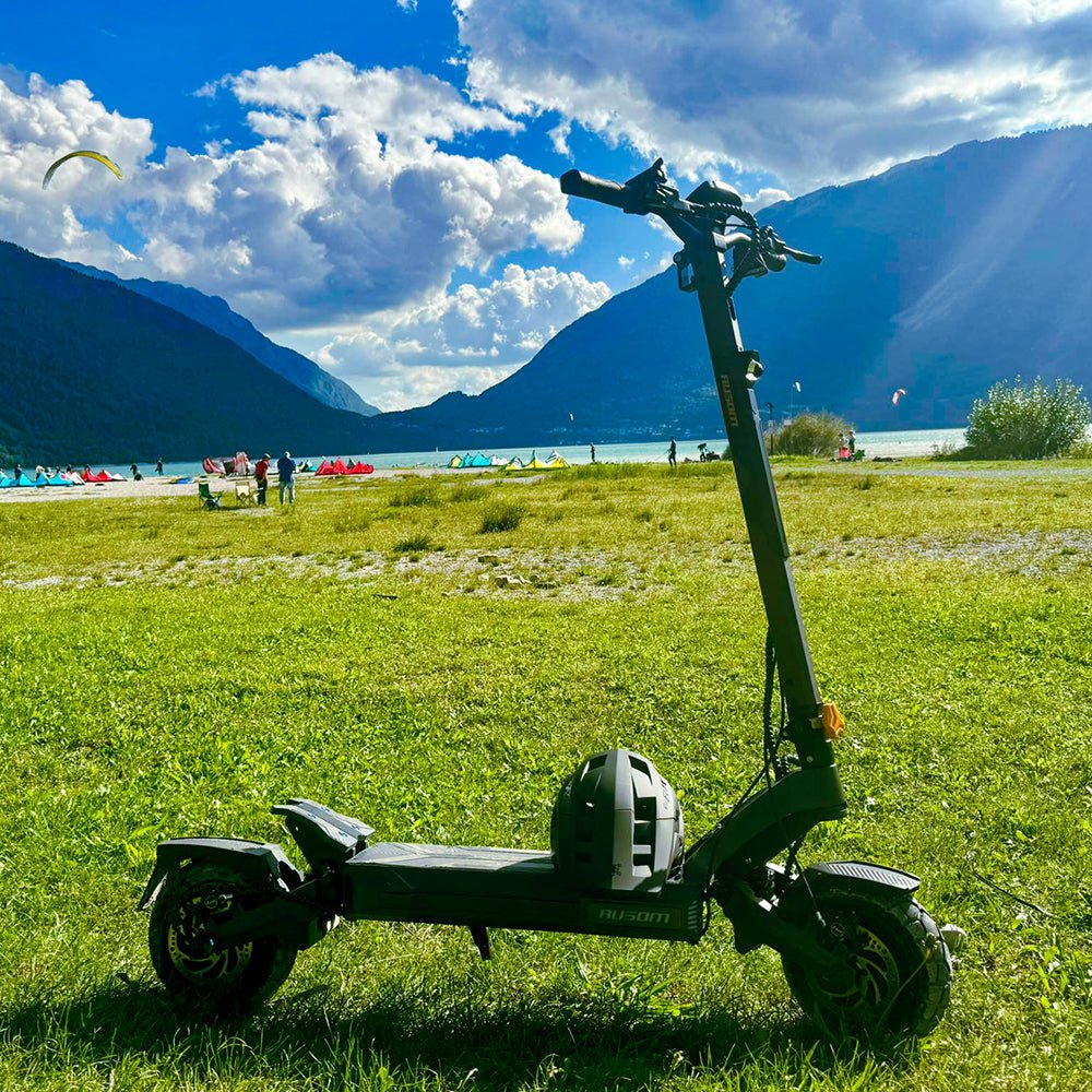Ausom DT2 Pro electric scooter standing on a lakeside meadow with mountains behind