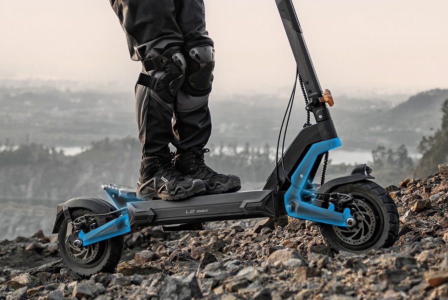Rider in knee armor standing on the Ausom L2 Max dual motor e-scooter on loose rock, showing the swingarm suspension