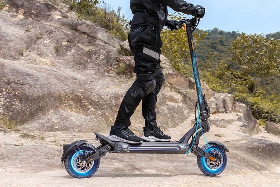 Rider standing on the Ausom SR1 electric scooter on a rocky trail showing both wheels and swingarms
