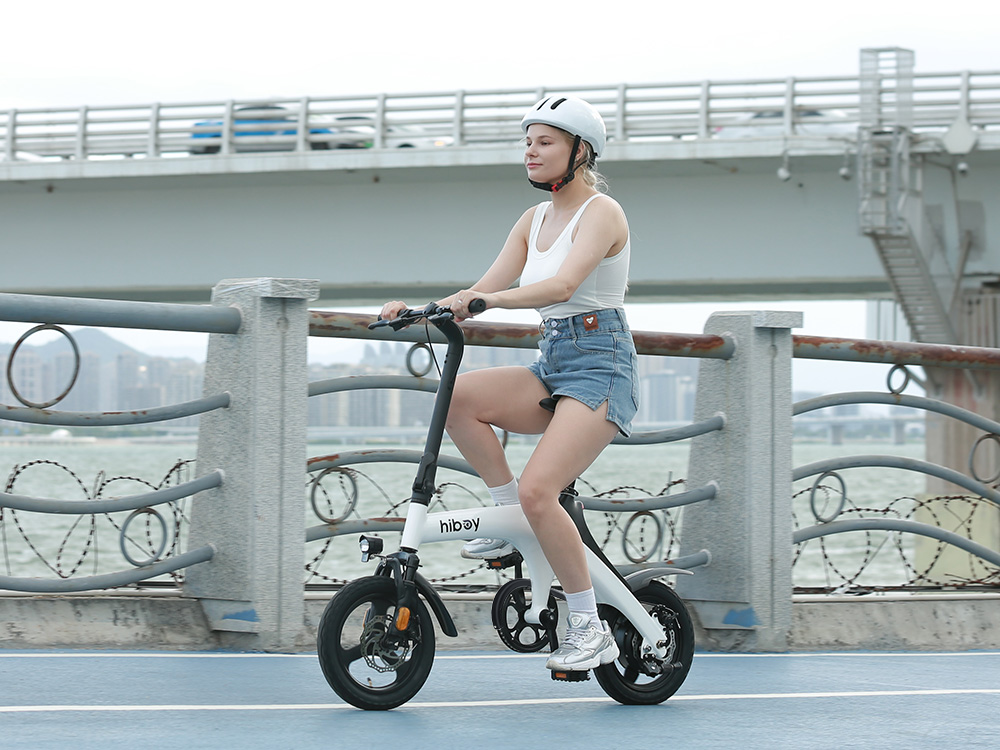 Rider in a white helmet pedaling the Hiboy C1 folding electric bike along a waterfront path