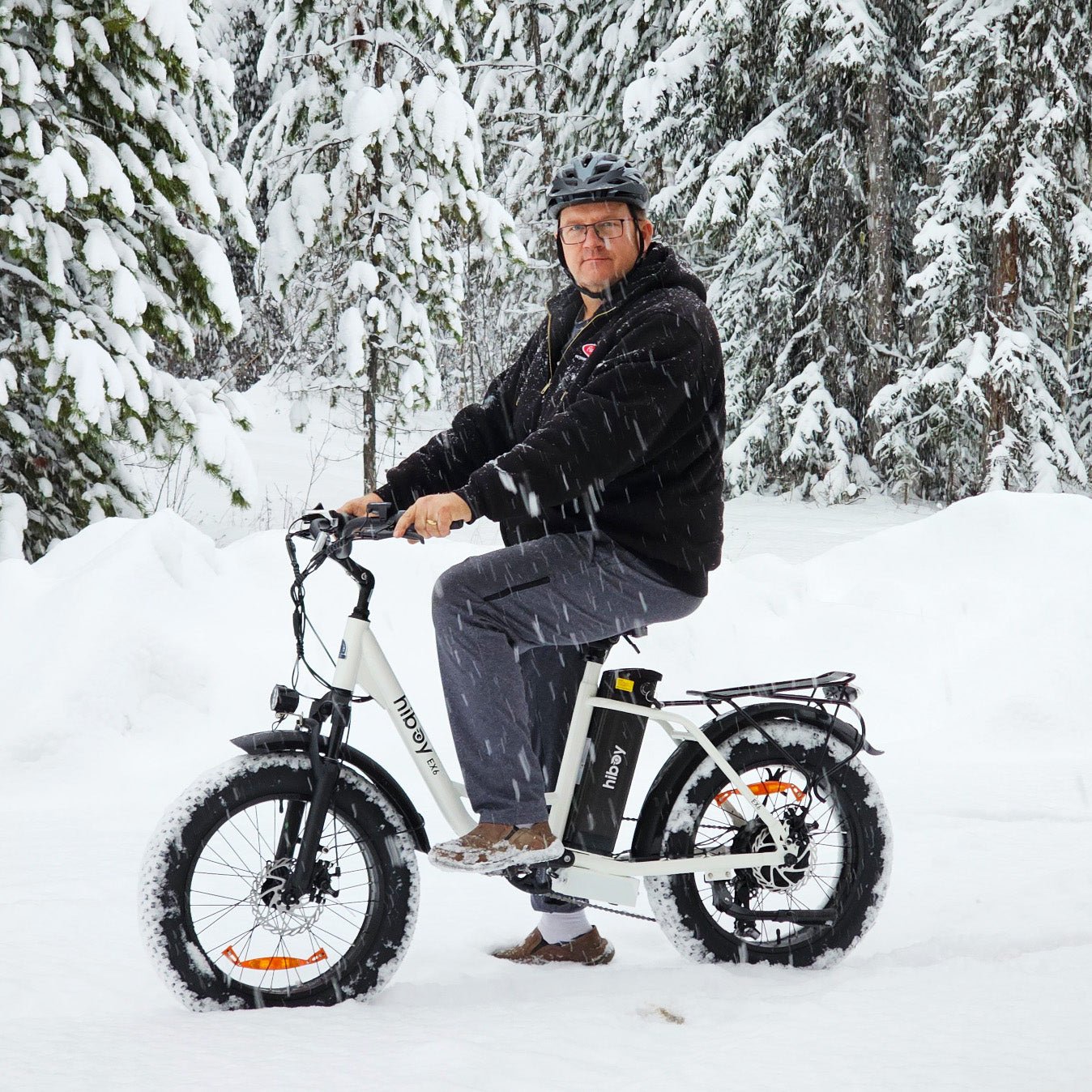 Rider on a white Hiboy EX6 electric bike in falling snow among pine trees
