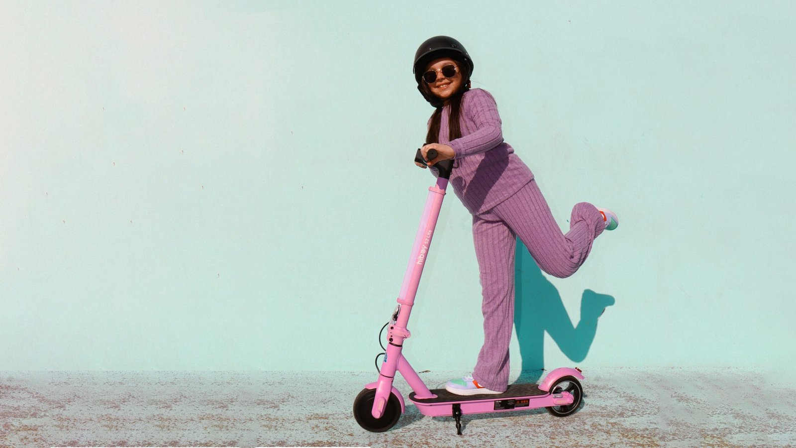 Young rider in a helmet kicking off on a pink Hiboy S2 Lite electric scooter against a turquoise wall