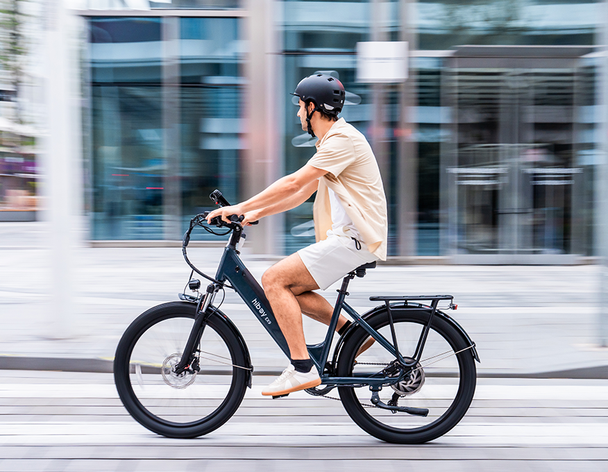 Rider on a navy Hiboy EX9 electric bike accelerating past glass office buildings