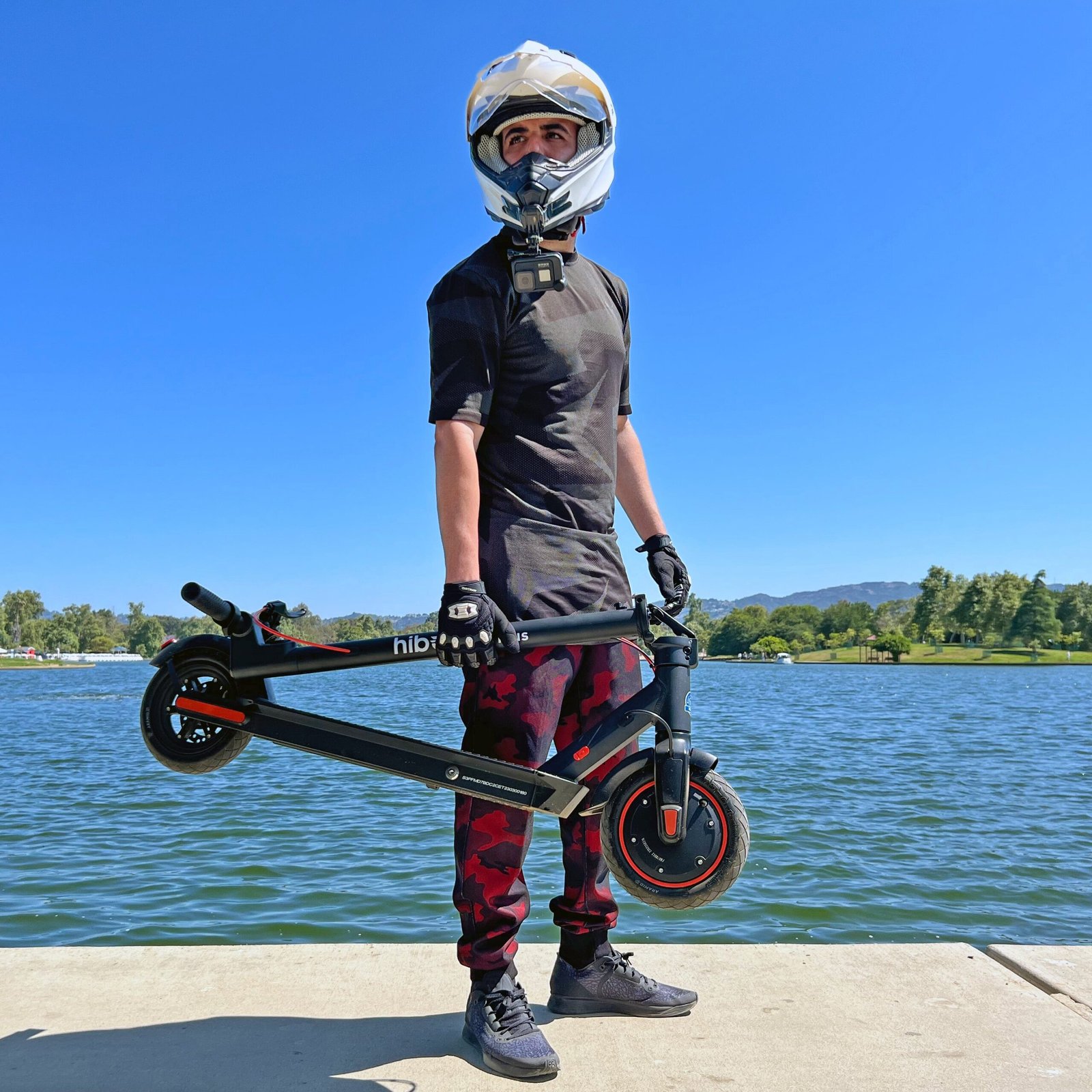 Rider in a helmet carrying the folded Hiboy S2R Plus electric scooter by a lake
