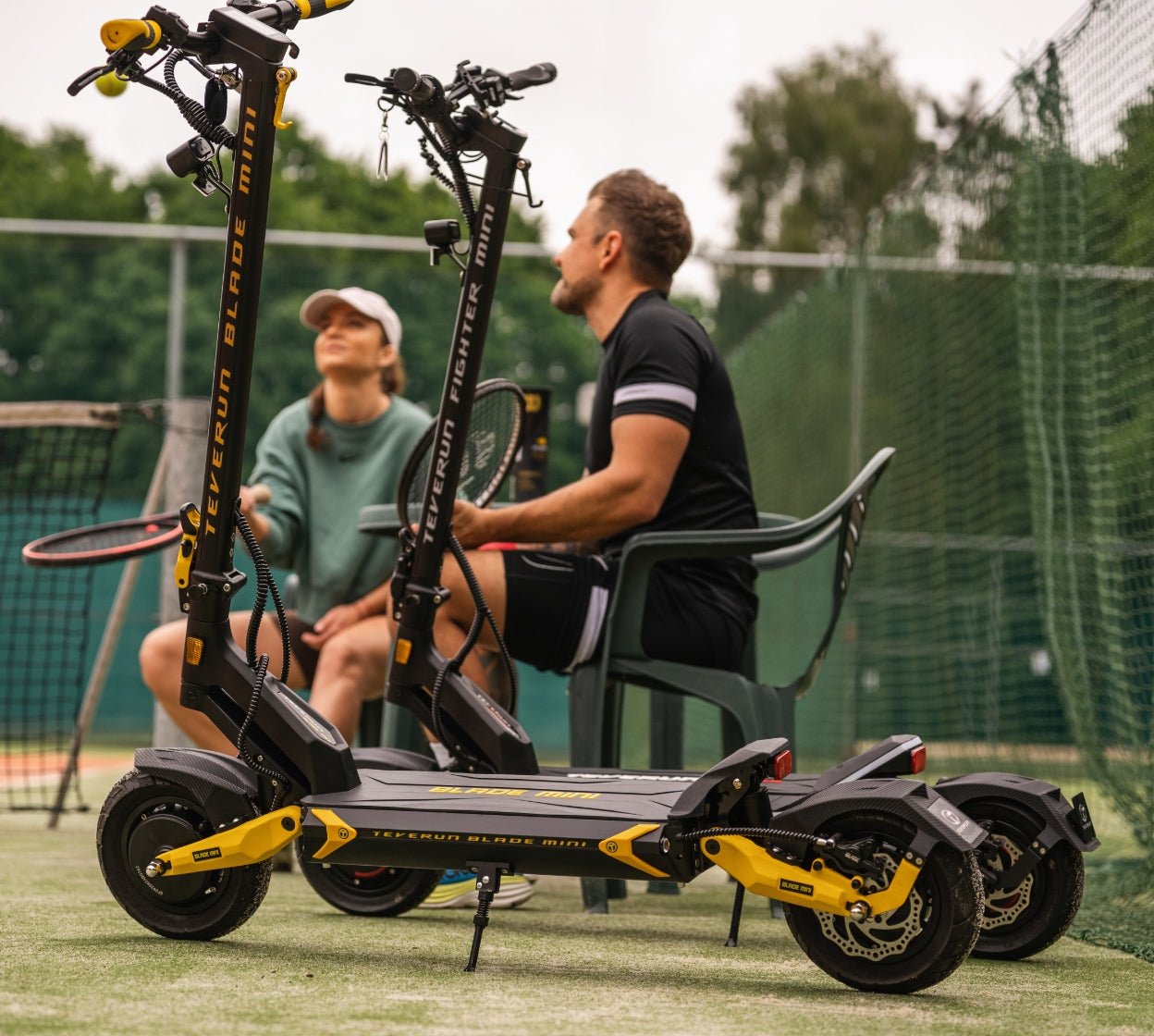 Two riders on the Teverun Blade Mini Ultra electric scooter beside a tennis court
