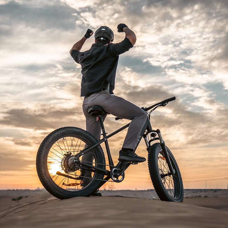 Rider celebrating with raised fists on a Hiboy P6 fat tire electric bike on a sand dune at sunset