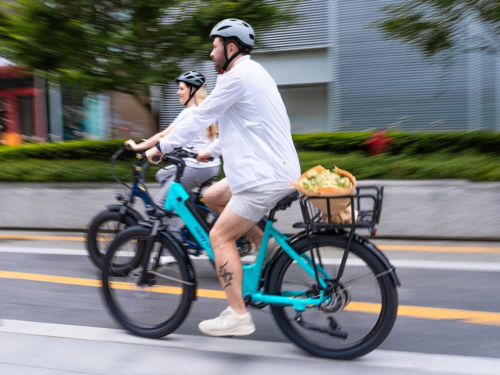 Two riders on teal and navy Hiboy EX9 electric bikes moving fast along a city street, flowers in the rear rack