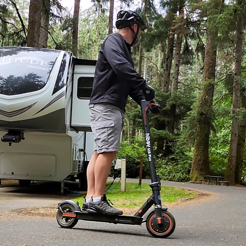 Rider on the Hiboy S2R Plus electric scooter at a forest campsite next to an RV